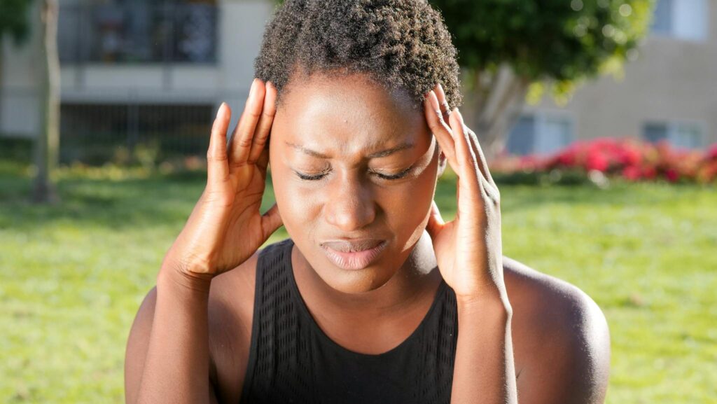 A woman holding her head in pain, indicating a severe headache outdoors in a sunny setting.