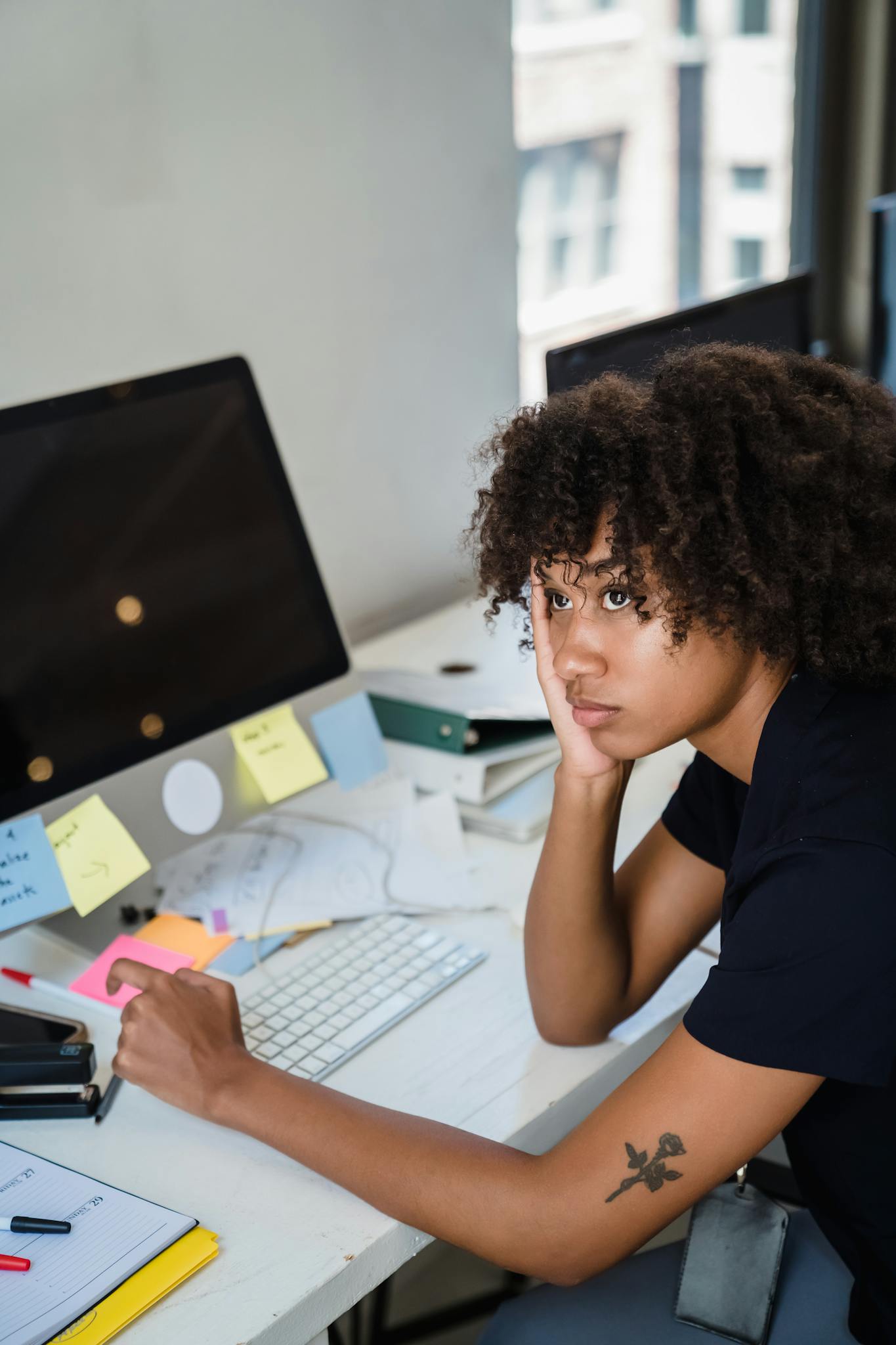 A woman looks tired while sitting at her work desk surrounded by computers and paperwork.